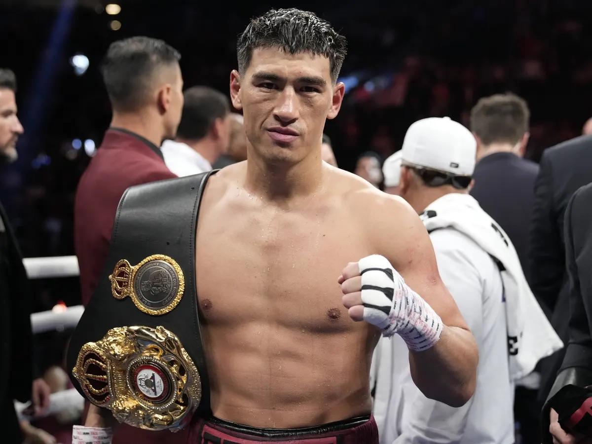 Dmitry Bivol posant avec sa ceinture WBA des poids mi-lourds après sa victoire, poing droit levé, torse nu, bandages aux mains, entouré de son staff dans l'arène — portrait du champion du monde WBA après avoir défendu son titre contre Eifert